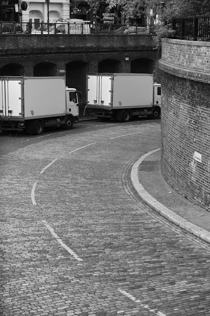 In this black and white image, three moving trucks with enclosed cargo areas are parked on a curved cobblestone street in an urban area, close to a brick wall and an arched underpass. The trucks are aligned along the pavement, with one positioned near the edge of the curve and the others further along the street. The scene shows an outdoor setting with a metal railing and some trees and buildings visible in the background above the brick wall. The street appears to be part of a residential or commercial district, and the trucks are likely in the process of a home relocation or furniture transport as part of a house removal service. The image captures the loading or parking phase of a professional moving process, possibly organized by Man with Van Kennington, within the context of ensuring efficient and careful packing and transporting of household items during a move.
