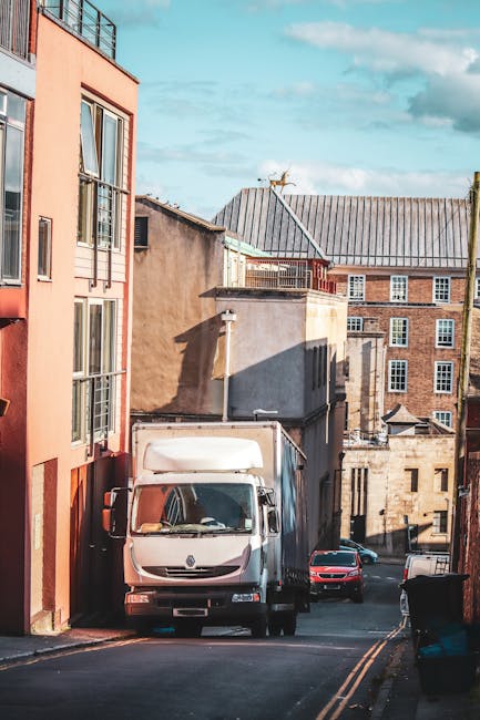 A white delivery van parked on a narrow urban street with a partially open cargo area at the rear, surrounded by multi-storey residential buildings made of brick and concrete. The van is positioned near the entrance of a building, suggesting it is involved in a home relocation or furniture transport process. Nearby, a red car is parked further down the street, and another vehicle is visible in the background. The street is paved with asphalt and has yellow double lines along the curb. The area is illuminated by daylight, with some shadows cast by the buildings, indicating clear weather. The image captures the loading or unloading scene typical of professional removals services such as those offered by Man with Van Kennington, highlighting the urban environment and logistics involved in house moves.