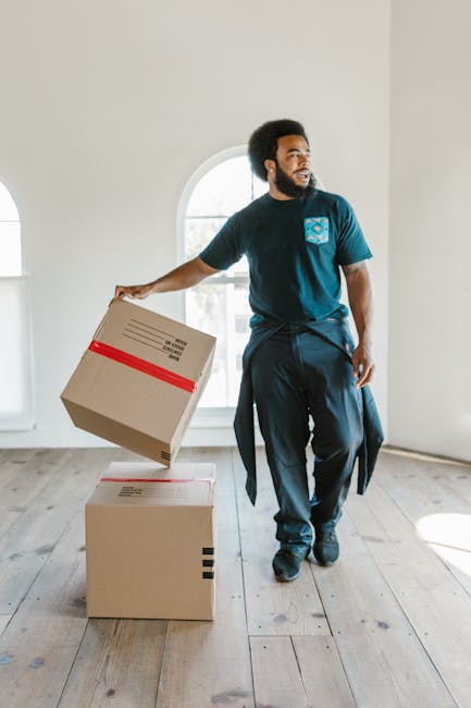 A man with curly hair, wearing a teal T-shirt and dark jeans, is seated on a wooden floor in a room with two large arched windows that allow natural daylight to illuminate the space. He is surrounded