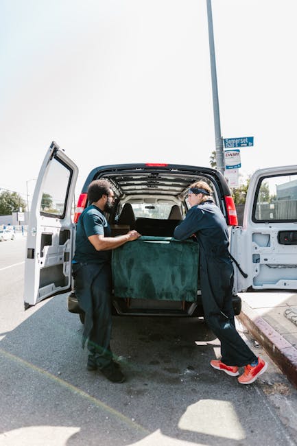 Two movers from Man with Van Kennington are engaged in a home relocation process, loading a large piece of furniture wrapped in dark green protective fabric into the open back of a black van parked on