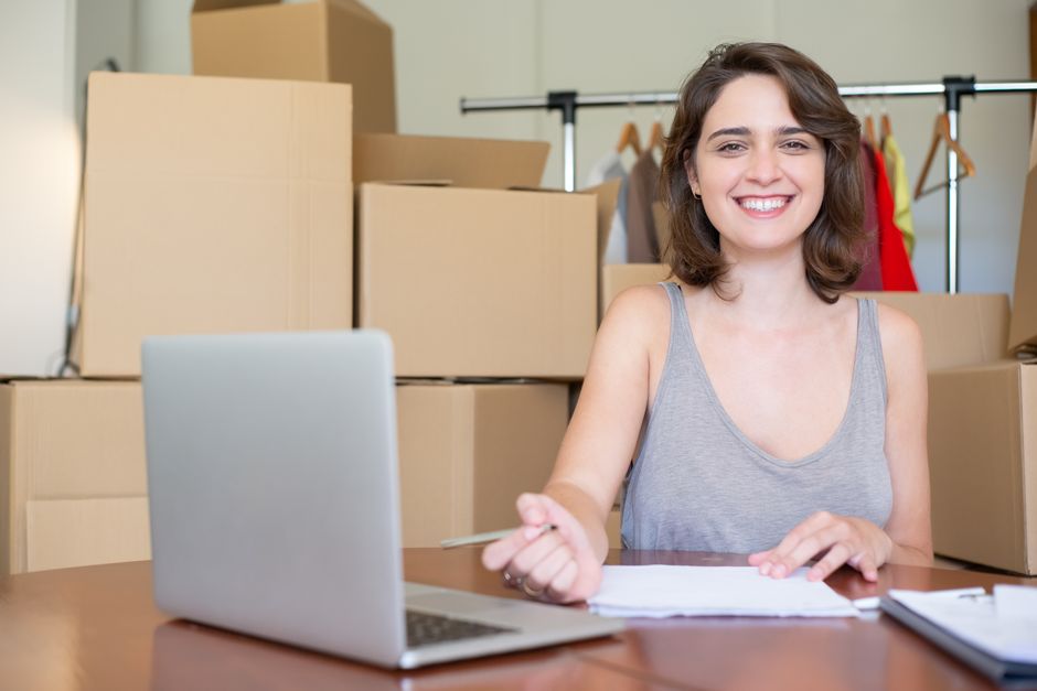 A woman with short, wavy brown hair and a bright smile is sitting at a desk in a room filled with moving boxes and packing materials. She is wearing a grey sleeveless top and holding a pen in her right hand, resting on a stack of papers on the desk. In front of her, there is an open silver laptop. Behind her, multiple large cardboard boxes are stacked, some sealed with packing tape, and others are partially open. To the right, a clothing rack with colorful garments is visible, indicating a house or office moving process. The lighting is natural, highlighting the tidy and organized environment during a packing and loading phase involved in a home relocation or furniture transport service. This scene illustrates a professional in the process of managing house removals, possibly coordinating logistics with Man with Van Kennington, as part of a small business move or local office relocation.