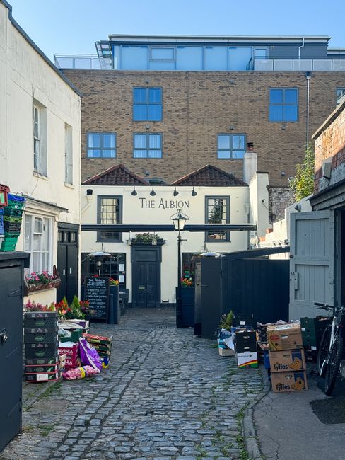 A narrow cobblestone alleyway leading to the entrance of The Albion, a small pub or restaurant housed in a white building with black window frames and signage, situated between grey fences and surrounded by various cardboard boxes and plastic crates filled with goods, indicating a moving or packing process related to house removals or furniture transport. The scene includes outdoor lighting fixtures, potted plants, and a clear sky above modern buildings in the background, suggesting a small-scale commercial or residential property with ongoing packing or loading activities, possibly coordinated by Man with Van Kennington during a home relocation or local house removals.