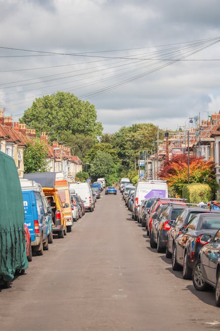 A residential street scene during daytime showing a line of parked vehicles on both sides of the narrow asphalt road, including vans, cars, and a covered vehicle, with some of the vans and cars displa
