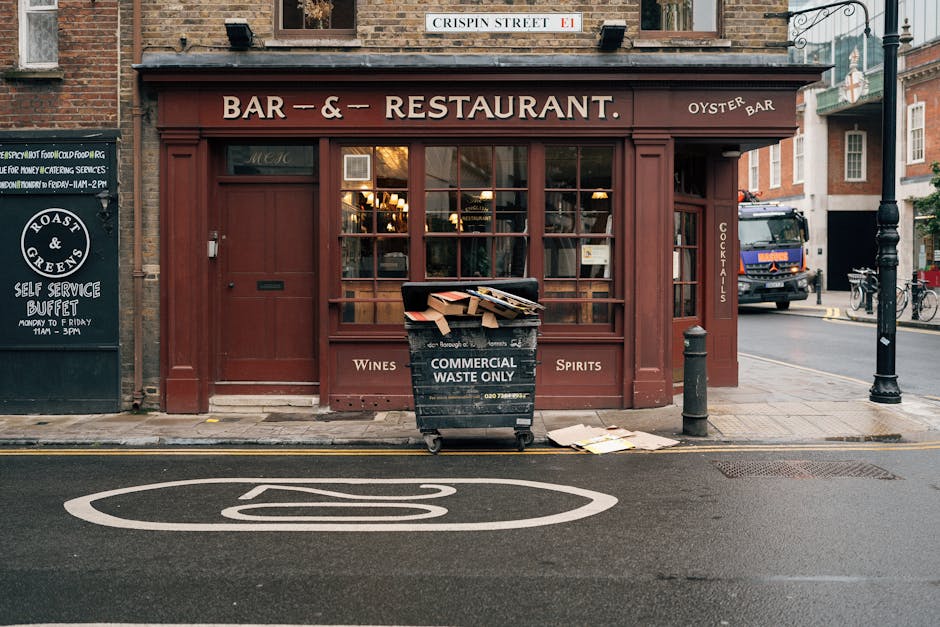 A red-brick building housing a bar and restaurant on Crispin Street, with large display windows revealing interior lighting. In front of the premises, there is a black commercial waste bin labeled 'COMMERCIAL WASTE ONLY' containing flattened cardboard boxes and discarded packing materials. The bin is positioned on the pavement near a black bollard and a small green post, with an open cardboard box and some loose packaging outside the bin. To the right, a large blue delivery truck is parked on the street, and bicycles are secured to a bike rack nearby. The scene depicts an urban environment suitable for house removals and furniture transport, with visible elements such as the waste collection area indicating ongoing logistics for moving or clearance activities. The image supports understanding of house removals, packing, and waste management in compliance with local regulations, aligned with [PAGE_TITLE] and the services provided by [COMPANY_NAME].