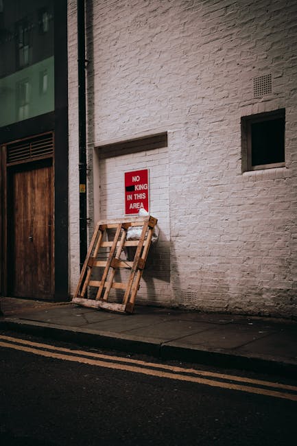 A red-brick building housing a bar and restaurant on Crispin Street, with large display windows revealing interior lighting. In front of the premises, there is a black commercial waste bin labeled 'COMMERCIAL WASTE ONLY' containing flattened cardboard boxes and discarded packing materials. The bin is positioned on the pavement near a black bollard and a small green post, with an open cardboard box and some loose packaging outside the bin. To the right, a large blue delivery truck is parked on the street, and bicycles are secured to a bike rack nearby. The scene depicts an urban environment suitable for house removals and furniture transport, with visible elements such as the waste collection area indicating ongoing logistics for moving or clearance activities. The image supports understanding of house removals, packing, and waste management in compliance with local regulations, aligned with [PAGE_TITLE] and the services provided by [COMPANY_NAME].