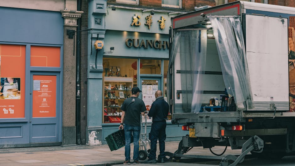 Two men are engaged in a home relocation process outside a storefront with the sign 'GUANGH.' One man, dressed in a black jacket and jeans, is holding a small dolly with a cardboard box, while the other, also in dark clothing, stands nearby. They are positioned on the pavement next to a moving truck with an open side panel, covered partially with a transparent plastic sheet, ready for loading or unloading furniture, packing containers, and other household items. The moving truck is parked close to the building’s entrance, with equipment such as straps and blankets likely on board to secure items during transport. The storefront features a large glass window displaying merchandise, and the building's exterior includes traditional elements like decorative columns and a concrete facade. This scene depicts a professional moving process, associated with house removals and furniture transport, supporting services like packing, loading, and home relocation, consistent with the context of 'Real removals costs in Kennington revealed' from Man with Van Kennington, as seen in the overall environment.