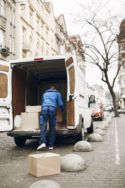 A man dressed in a blue jacket and dark trousers is loading or unloading cardboard boxes into the back of a white removal van parked on a city street. The van's rear doors are open, revealing an interior with visible wooden paneling and large packing boxes. A cardboard box rests on the pavement near his feet. The street is lined with residential buildings featuring ornate facades, and trees with bare branches are visible in the background. Several parked cars are along the curb, and large, rounded cobblestone sculptures separate the parking area from the sidewalk. The scene is set during daylight, with natural light illuminating the environment, reflecting typical furniture transport and packing activities associated with home relocation services, as offered by Man with Van Kennington, during a house removals project.