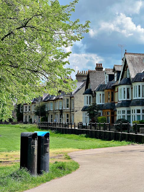 A row of residential houses with bay windows and pitched roofs, situated along a street in Kennington, with a large leafy green tree in the foreground and a partly cloudy blue sky overhead. In front of the houses, a grassy area with a small paved pathway is visible, alongside black metal fencing separating the greenery from the sidewalk. Two black wheelie bins are positioned on the grass near the path, with one bin displaying a recycling symbol. The scene depicts a quiet neighborhood environment. The image supports the context of home relocation and moving services, as provided by Man with Van Kennington, by illustrating the local residential setting where furniture transport and packing may occur.