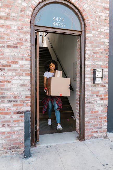 A young woman walking through a narrow brick building entrance with a rounded archway, carrying a large cardboard box with red tape, and several smaller boxes stacked in her arms. She is dressed in casual clothing, including a white t-shirt, blue jeans, and white sneakers, with a red and black checkered shirt tied around her waist. Behind her, a staircase with dark steps and a metal handrail is visible inside the building, indicating an ongoing home relocation or moving process. Outside, the pavement and sidewalk are visible, with a doorbell or intercom panel mounted on the brick wall to the right of the entrance. The scene captures a moment during packing and furniture transport, with natural lighting highlighting the details of the moving activity as part of a house removal service offered by Man with Van Kennington, supporting efficient and careful furniture transport near Oval Station.