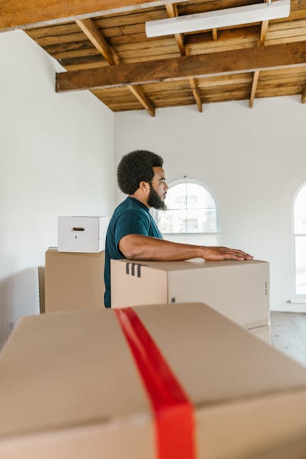 A man with curly hair and medium skin tone, wearing a teal t-shirt and dark pants, sits on a wooden floor inside a room with large arched windows. The room is illuminated by natural light, and several cardboard boxes of various sizes, sealed with red tape, are stacked behind him, indicating a home relocation or packing process. A tall potted plant with broad green leaves stands to the right of the man next to the window. The scene captures a moment during packing or moving preparations, with the boxes ready for transportation. The overall environment suggests an indoor setting during the moving process managed by a professional removals service, such as Man with Van Kennington, supporting house removals and furniture transport.