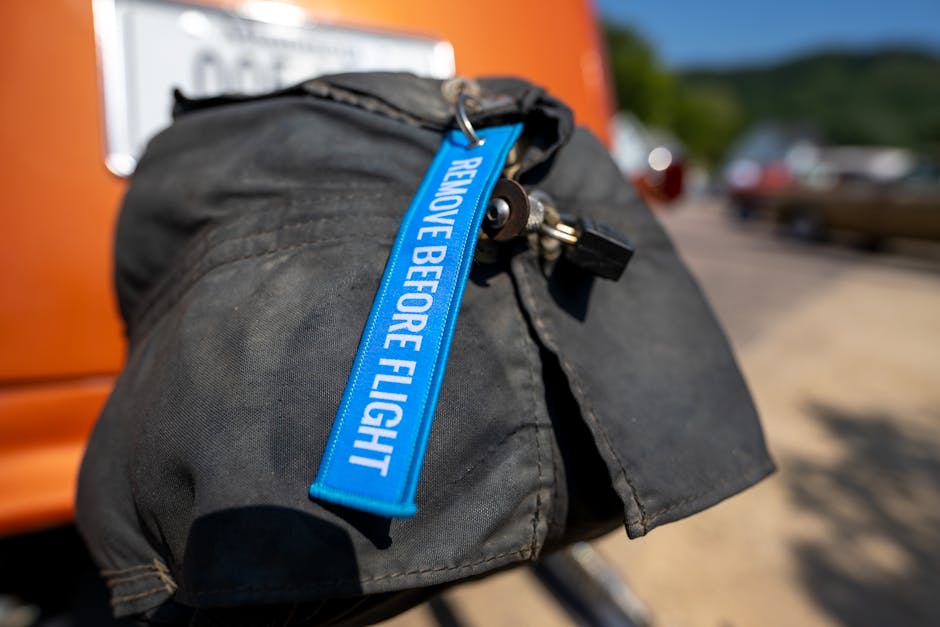 Close-up view of a black nylon bag secured with a blue strap that has the words 'REMOVE BEFORE FLIGHT' printed on it. The bag, likely used for packing belongings during a house removal, is placed on a trolley or hand truck in an outdoor setting. Behind the bag, the background features an orange van with blurred elements of a narrow residential street, including parked cars and trees, indicating a home relocation process in progress. The lighting suggests daytime with clear weather, and the scene captures the preparation stage of loading items into a vehicle by Man with Van Kennington, a professional removals service specializing in domestic moves. The image emphasizes careful packing, loading procedures, and the logistics involved in furniture and box transport for a house move, especially in tight street conditions often encountered in London neighborhoods like Kennington.