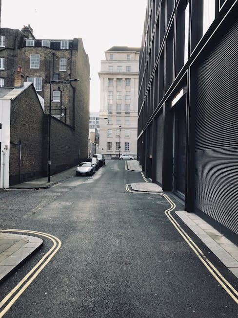 A narrow residential street in Kennington during late afternoon or early evening, with parked cars lining both sides of the road. The street is shaded by tall trees with dense green foliage, creating dappled sunlight on the asphalt. In the distance, a white van belonging to Man with Van Kennington is partially visible, positioned near the curb for a house removal or furniture transport operation. The van is aligned with the pavement, ready for loading or unloading, with furniture and boxes possibly inside or being moved outside. The surrounding environment includes a wooden fence, a bus stop sign, and the rooftops of houses partly obscured by the trees. The scene captures the typical setting for a home relocation in a quiet urban area, emphasizing careful vehicle positioning for parking and safe loading in a narrow street context, consistent with a professional removals service.